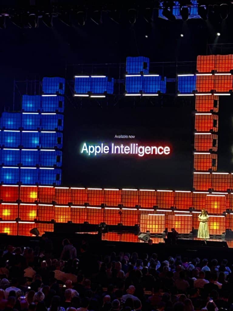Cette photo montre une scène lors d'une conférence, avec une grande inscription lumineuse affichant Apple Intelligence au centre de l'écran principal. La scène est entourée de panneaux LED colorés en bleu et rouge, créant une ambiance technologique et dynamique. Une personne se tient sur scène devant un public nombreux, semblant présenter ou annoncer le produit ou service Apple Intelligence. L'éclairage et la disposition visuelle mettent en valeur l'aspect innovant de l'événement.