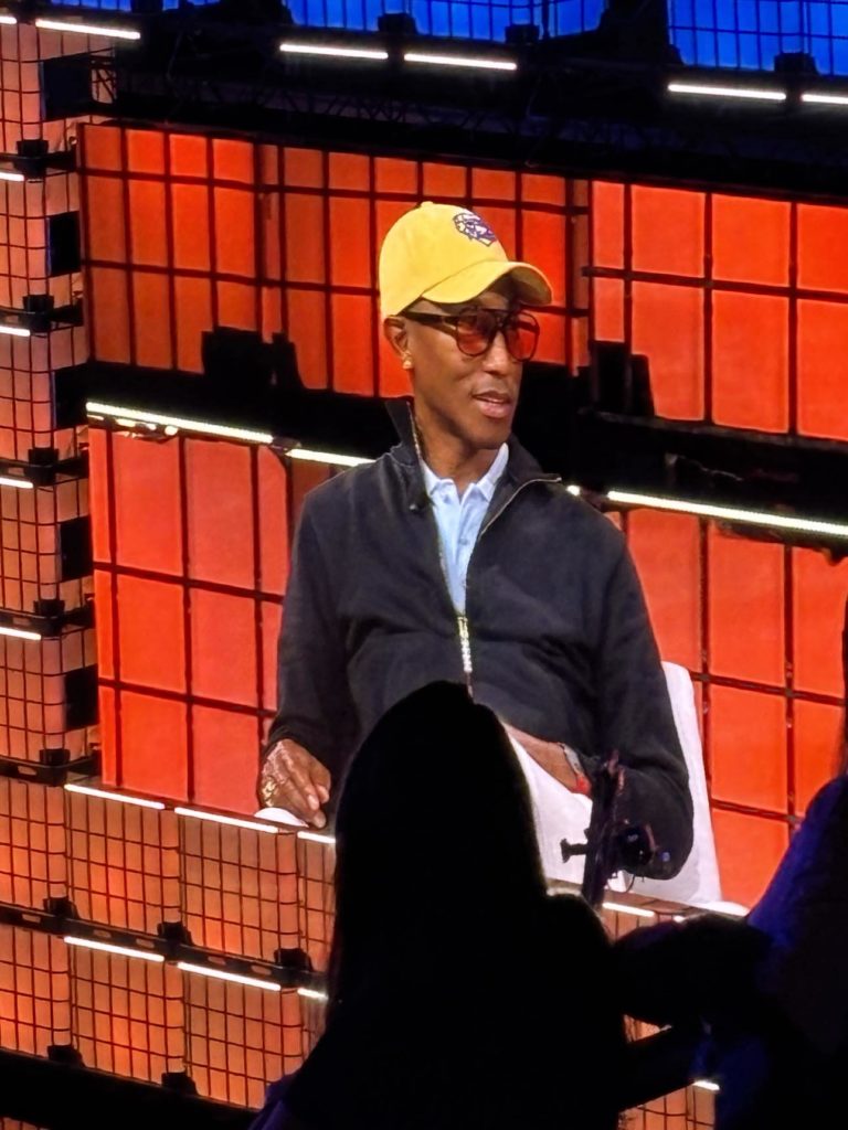 L'image montre Pharrell Williams, identifiable par sa casquette jaune et son style distinctif, assis sur une scène lors d'un événement. Il porte des lunettes de soleil et une tenue décontractée. En arrière-plan, des panneaux lumineux aux teintes rouges et orange forment un décor moderne et technologique. Une silhouette au premier plan, probablement celle d'une personne dans le public ou d'un caméraman, renforce l'idée d'une conférence ou d'une présentation.