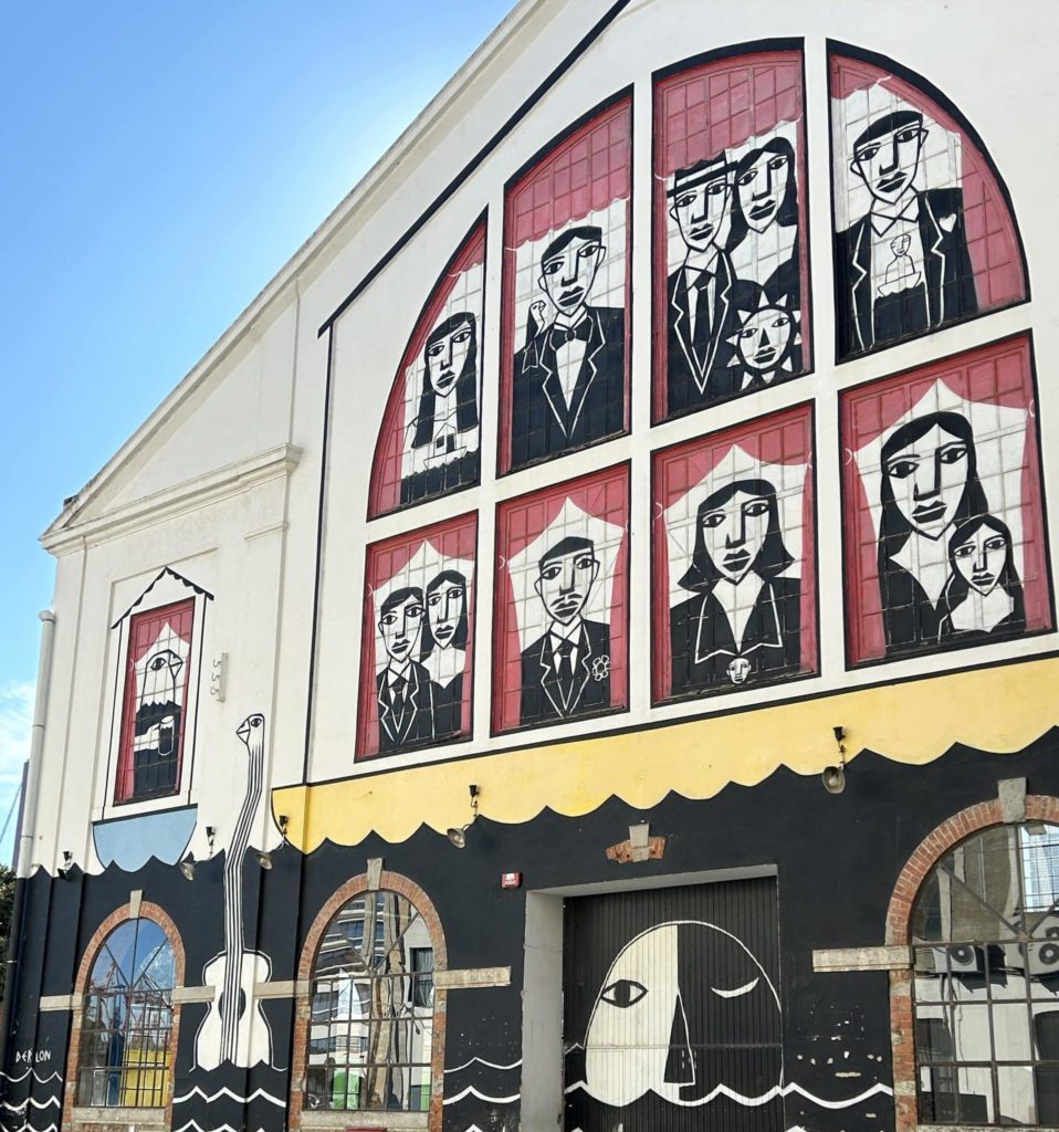 L'image montre une fresque murale peinte sur la façade d'un bâtiment. La partie supérieure de la fresque représente des visages stylisés dans des cadres arqués rouges, rappelant des fenêtres ou des vitraux. Les personnages ont un style graphique en noir et blanc, avec des traits géométriques et des expressions sobres. La partie inférieure de la fresque inclut des éléments marins, comme une vague et une figure abstraite avec un visage semi-circulaire. Le design utilise des couleurs contrastées, notamment le noir, le blanc, le rouge et le jaune, créant une composition artistique marquante.