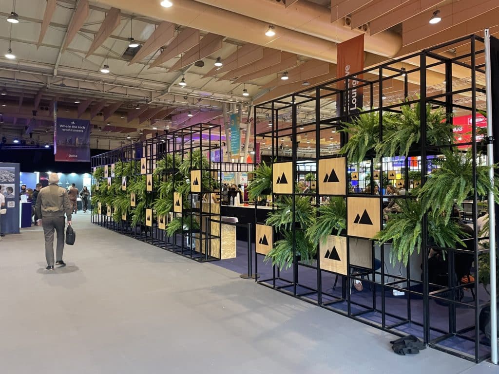 La photo montre un espace d'exposition intérieur avec une structure métallique composée de cadres noirs, ornée de plantes vertes et de panneaux en bois affichant un logo représentant deux triangles noirs sur un fond clair. L'ensemble crée une ambiance naturelle et moderne. À l'arrière-plan, on distingue des stands et des zones d'accueil typiques d'un événement professionnel ou d'une conférence, avec des panneaux suspendus indiquant différentes zones comme le lounge. Des visiteurs déambulent dans les allées, explorant les installations. L'éclairage est chaleureux, mettant en valeur l'espace et les éléments visuels.