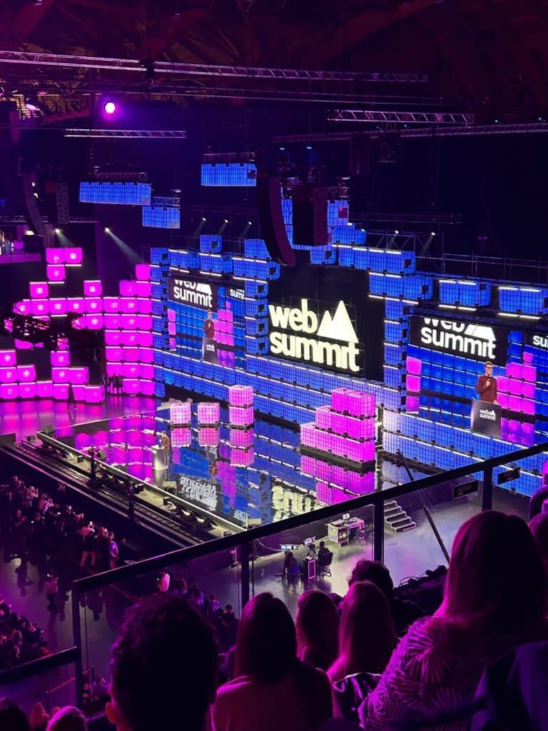 Cette image montre une vue d'ensemble de la scène principale du Web Summit. La scène est illuminée par des panneaux LED colorés en violet, bleu et rose, créant une atmosphère vibrante et technologique. Le logo du Web Summit est bien visible au centre, affiché en blanc sur fond noir. Le public est installé dans les gradins au premier plan, observant l'événement. La disposition et l'éclairage donnent une impression d'énergie et d'ampleur, typique d'un grand rassemblement technologique.