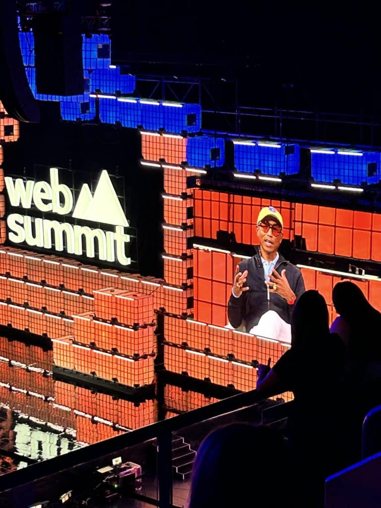 Cette image montre Pharrell Williams sur scène lors du Web Summit, reconnaissable à sa casquette jaune et son style décontracté. Il semble s'exprimer, accompagné de gestes pour appuyer son discours. L'arrière-plan est constitué de panneaux lumineux orange et rouges avec le logo du Web Summit affiché en blanc sur la gauche. Au premier plan, des silhouettes de spectateurs ajoutent une dimension immersive, évoquant un événement dynamique et technologique.