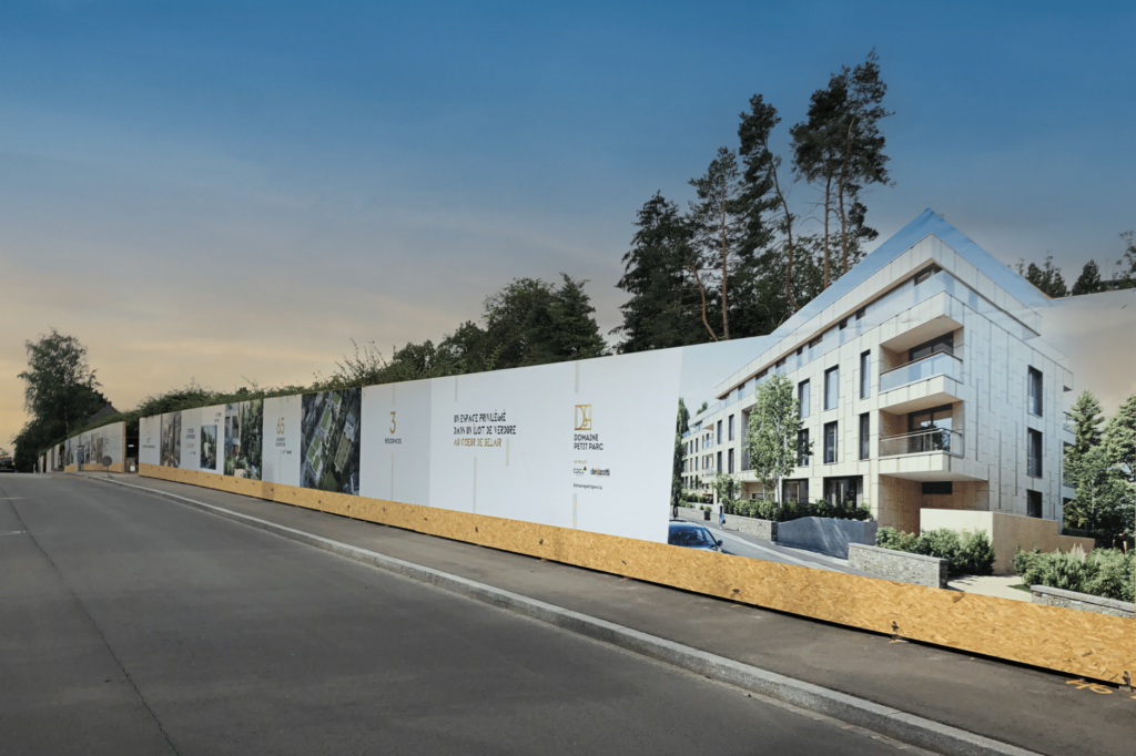 Bâche de chantier le long d'une route affichant le projet immobilier Domaine Petit Parc avec des visuels d'immeubles modernes, des textes promotionnels et des logos sur fond blanc, entourée de végétation et d'arbres.