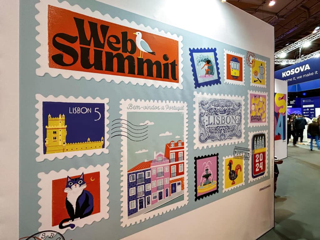 L'image montre un panneau décoratif présenté au Web Summit, avec un design inspiré de timbres-poste. Chaque timbre illustre des éléments emblématiques de Lisbonne et du Portugal, comme la Tour de Belém, un tramway jaune, des maisons colorées, un coq de Barcelos, et des motifs traditionnels portugais. Le logo du Web Summit est placé en haut, avec une mouette posée sur le mot "Summit". Le texte "Bem-vindos a Portugal" souhaite la bienvenue aux visiteurs. L'ensemble est lumineux et coloré, créant une ambiance accueillante et culturelle.