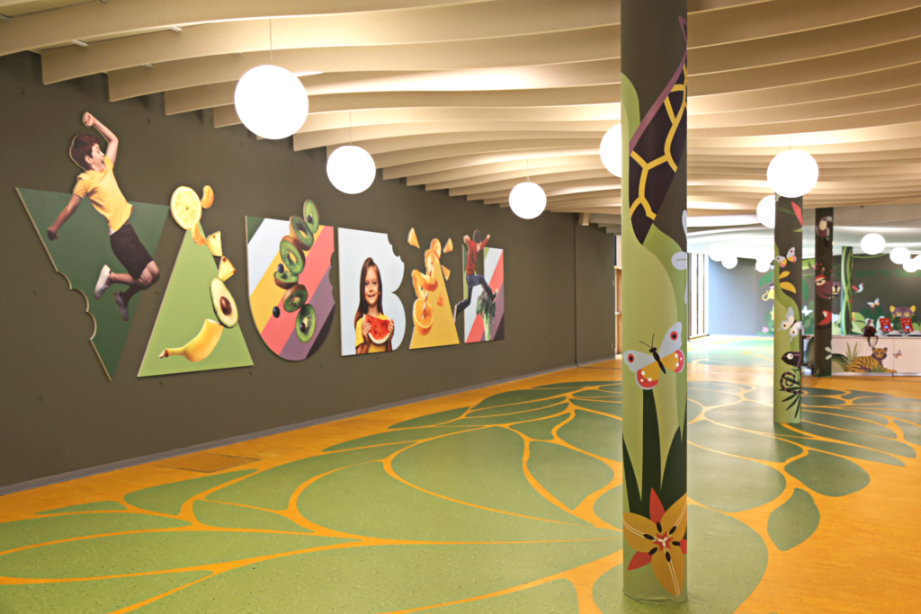 L'image montre un espace intérieur coloré et lumineux, probablement destiné aux enfants, avec un sol en motifs verts et jaunes évoquant des feuilles ou des plantes. Les murs sont décorés avec des panneaux en relief représentant des formes géométriques et des images de fruits, légumes, et enfants joyeux. Les colonnes sont illustrées avec des dessins de papillons, de fleurs et d'éléments naturels. Le plafond est orné de structures ondulées et des luminaires sphériques suspendus, ajoutant une ambiance douce et accueillante. L'ensemble crée un environnement ludique et éducatif centré sur la nature et la santé.