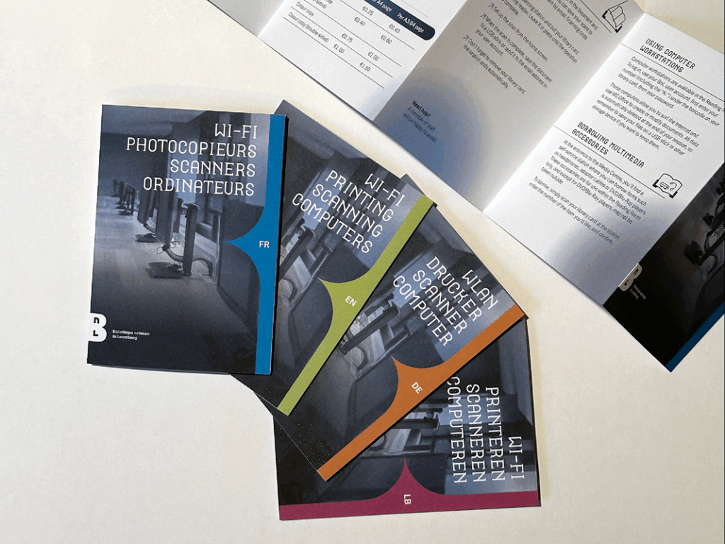 Quatre brochures multilingues posées sur une table, publiées par la Bibliothèque nationale du Luxembourg. Chaque brochure présente les services informatiques disponibles (Wi-Fi, photocopieurs, scanners, ordinateurs) en une langue différente : français (bleu), anglais (vert), allemand (orange) et luxembourgeois (rose). En arrière-plan, une brochure ouverte montre des explications détaillées sur l’utilisation des ordinateurs et le prêt d’accessoires multimédias.
