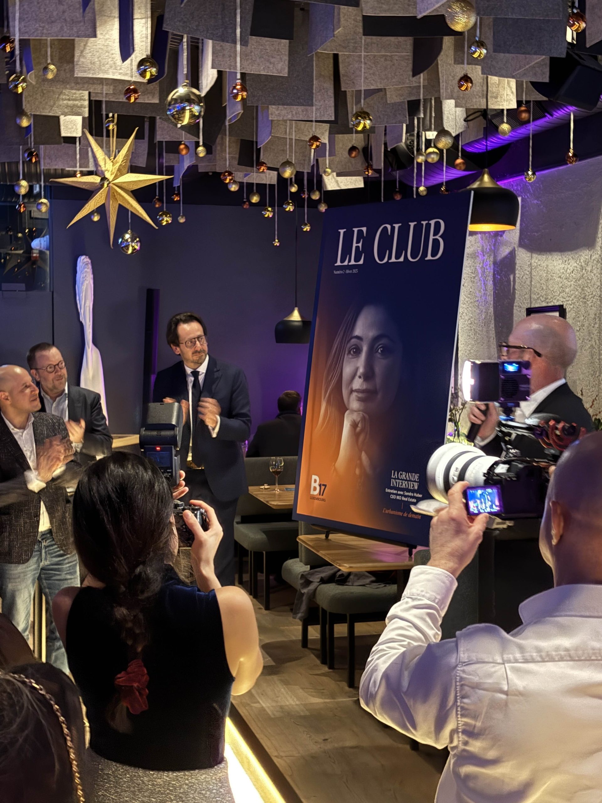 Scène dans un bar ou un lounge décoré de suspensions et boules dorées. Un groupe de personnes, dont un homme en costume applaudissant, entoure un grand panneau présentant la couverture du magazine Le Club. Plusieurs photographes prennent des clichés du magazine exposé