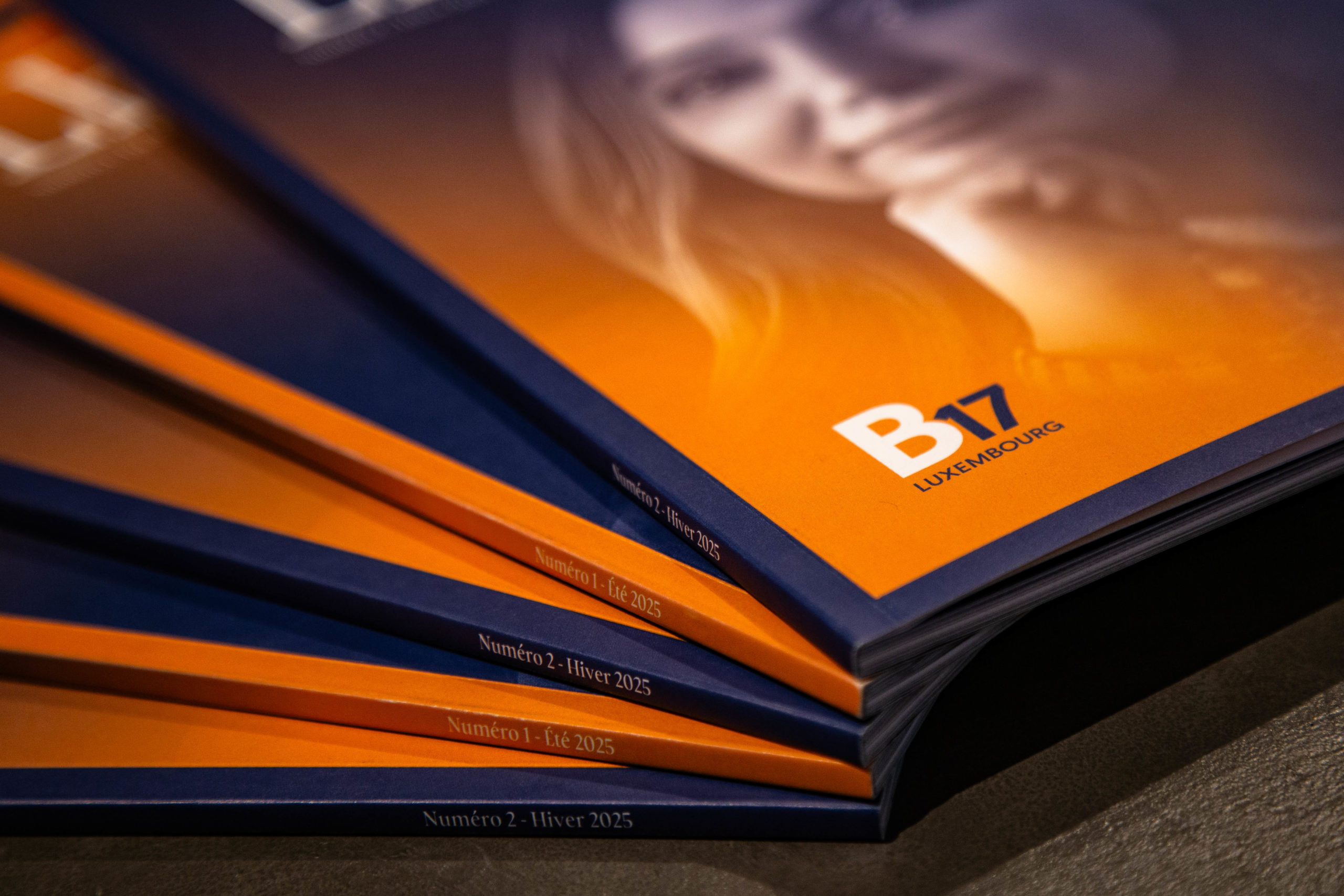 Gros plan sur une pile de magazines édités par le B17 Luxembourg, mettant en valeur leurs tranches et couvertures aux couleurs orange et bleu marine.