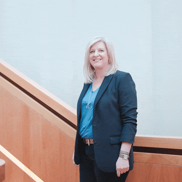 Femme blonde souriante en veste de tailleur sombre et haut bleu, posant dans un escalier en bois.