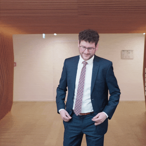 : Portrait d'un homme portant des lunettes et un costume bleu foncé à côté d'une paroi en lattes de bois.