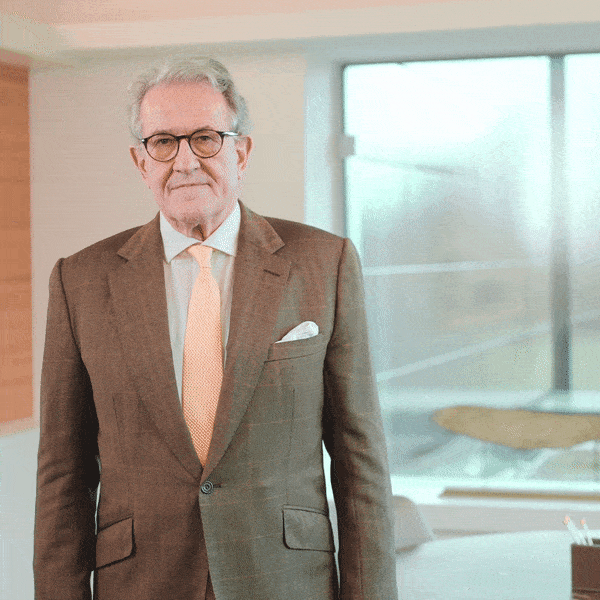 Portrait d'un homme senior portant des lunettes, un costume marron et une cravate orange dans un bureau lumineux.