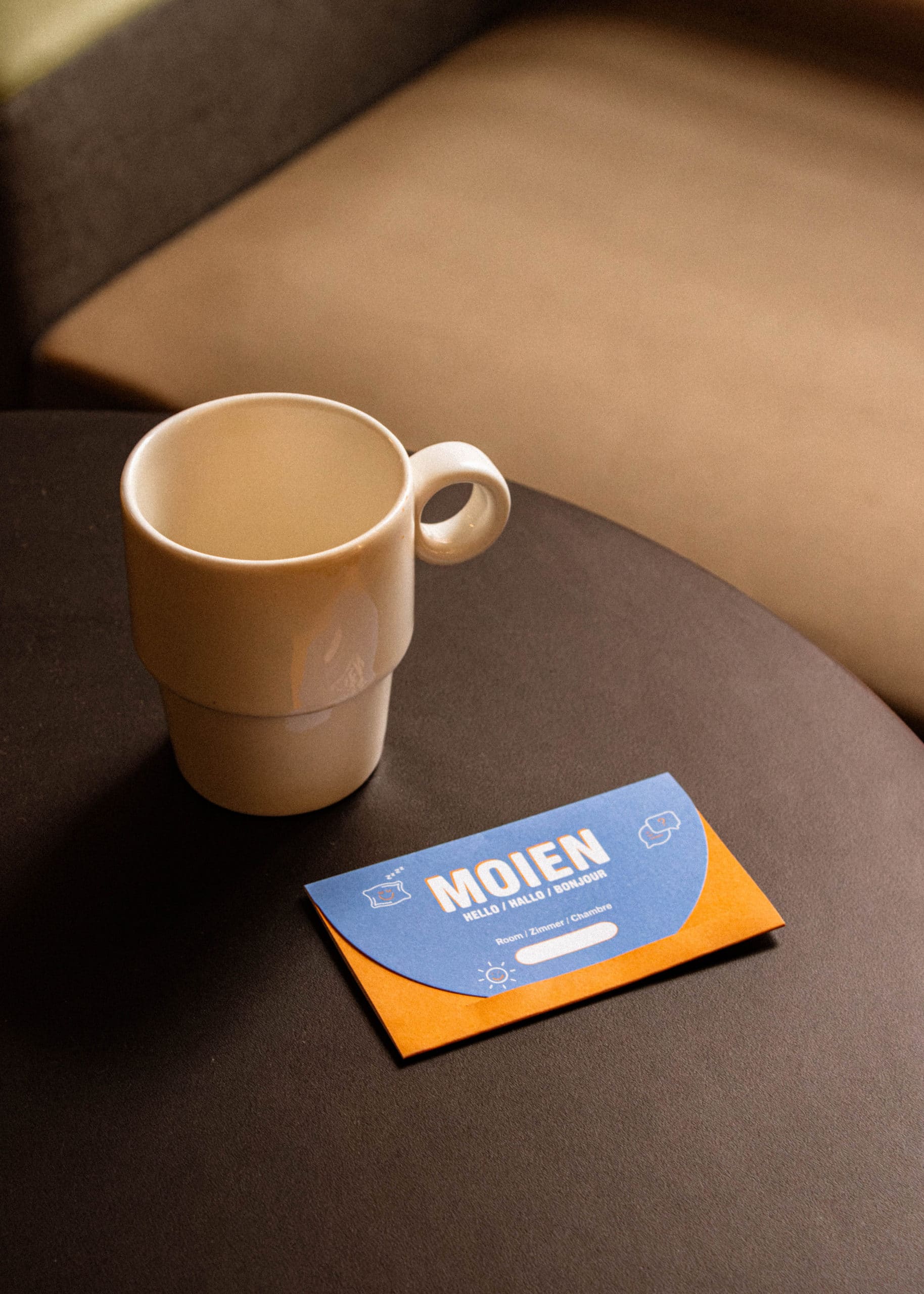 Pochette bleue et orange pour clé de chambre indiquant "Moien", posée à côté d'une tasse blanche.