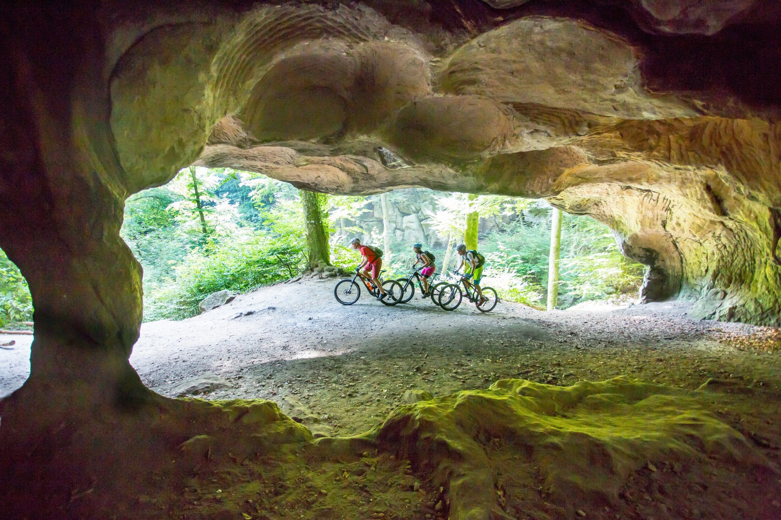 Groupe de cyclistes en VTT explorant les formations rocheuses et la forêt de la région du Mullerthal.