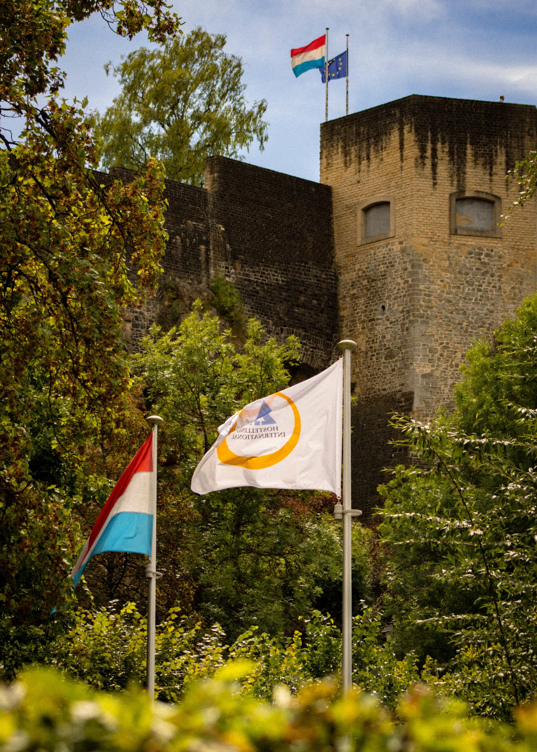 Cadre extérieur de l'auberge de jeunesse de Luxembourg située au pied des fortifications historiques, avec les drapeaux luxembourgeois et Hostelling International au premier plan.