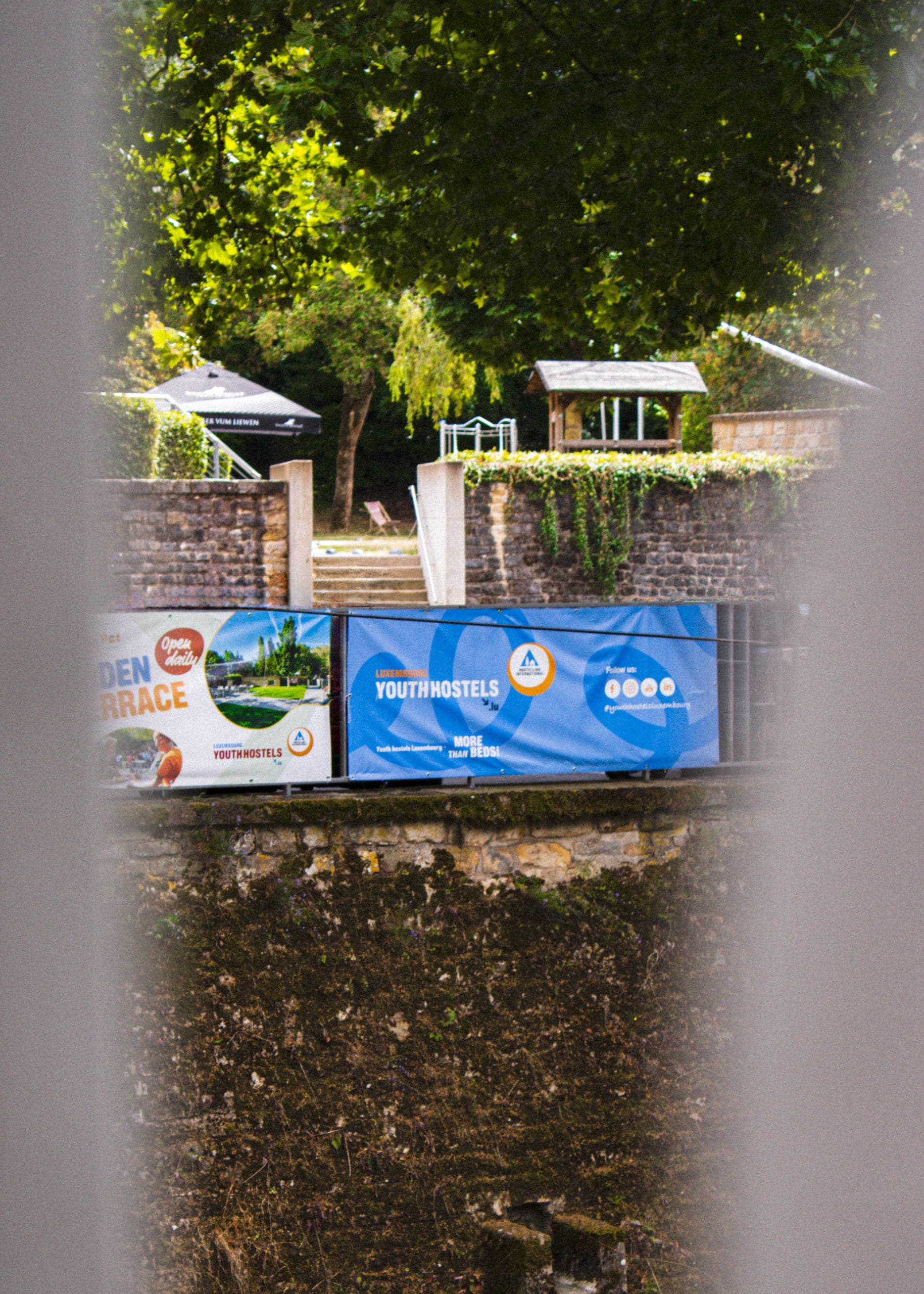 Bannières publicitaires Luxembourg Youth Hostels annonçant la terrasse ouverte sur un mur en pierre, avec un aperçu du jardin et des escaliers de l'auberge en arrière-plan.