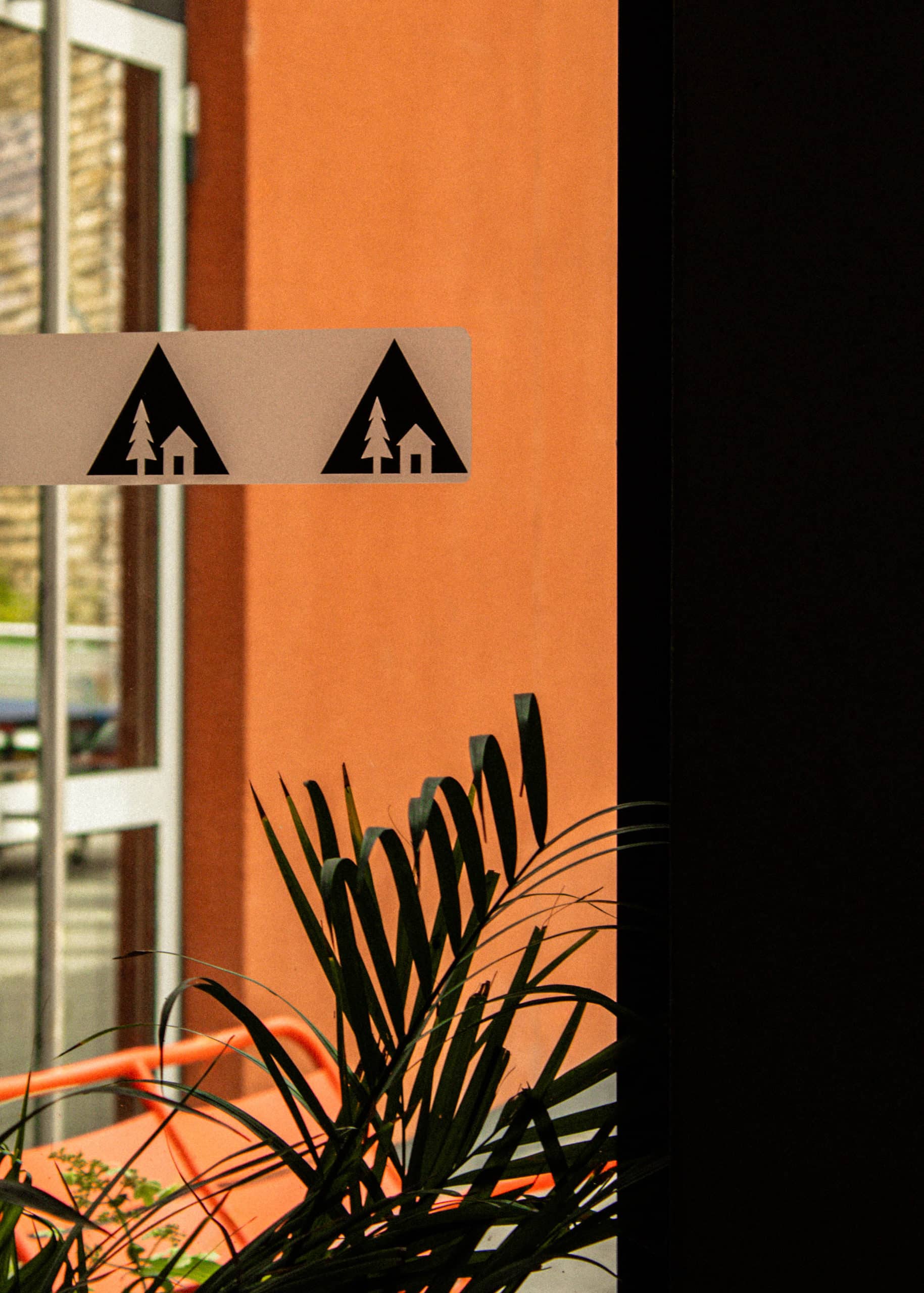 Signalétique sur la vitrine orange d'une auberge de jeunesse Luxembourg Youth Hostels, avec des pictogrammes de sapin et de maison, partiellement masquée par une plante verte.