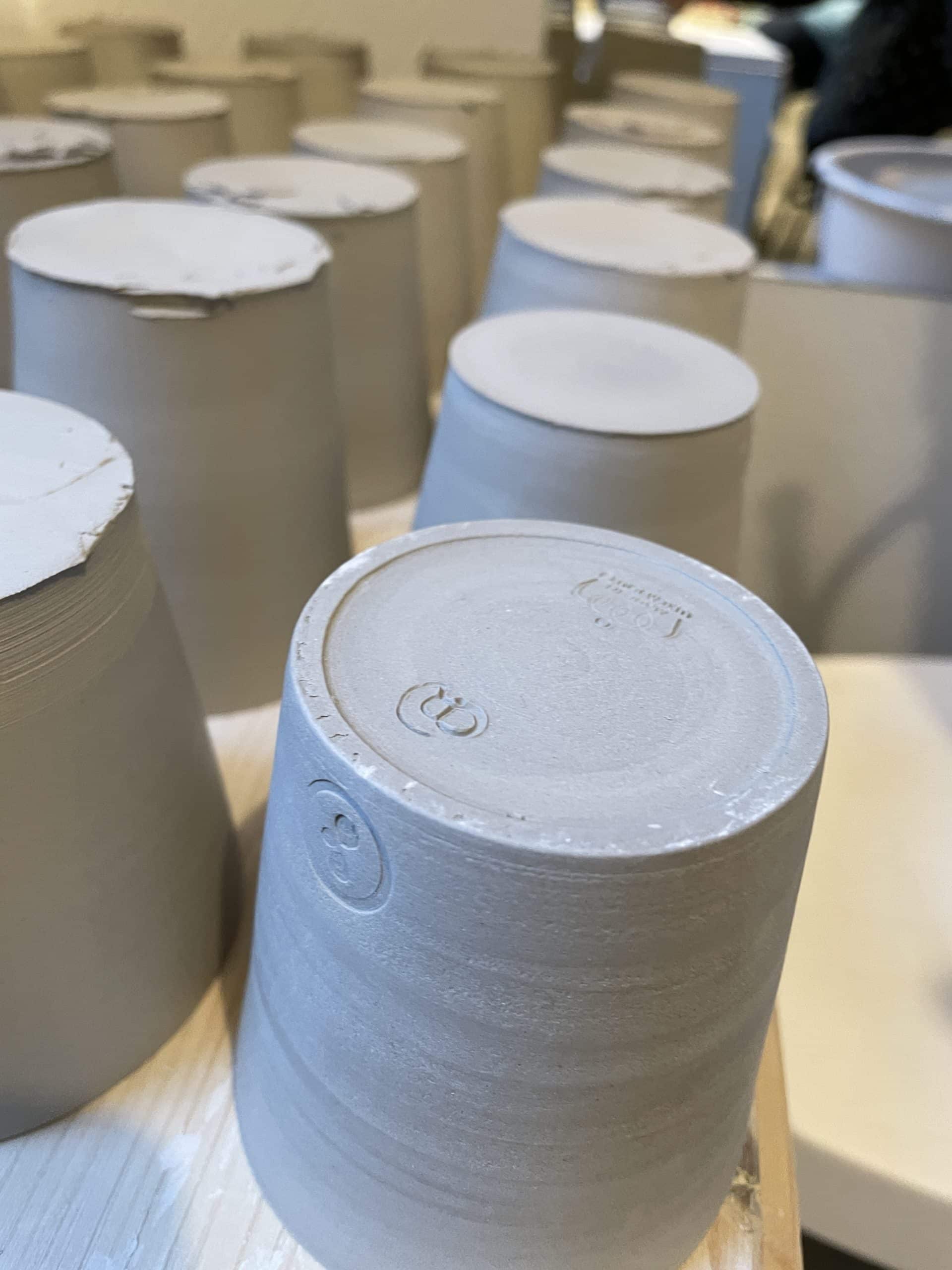 Pots en argile grise crue séchant à l'envers sur un plan de travail en bois, avec un poinçon d'artisan gravé sur le fond.