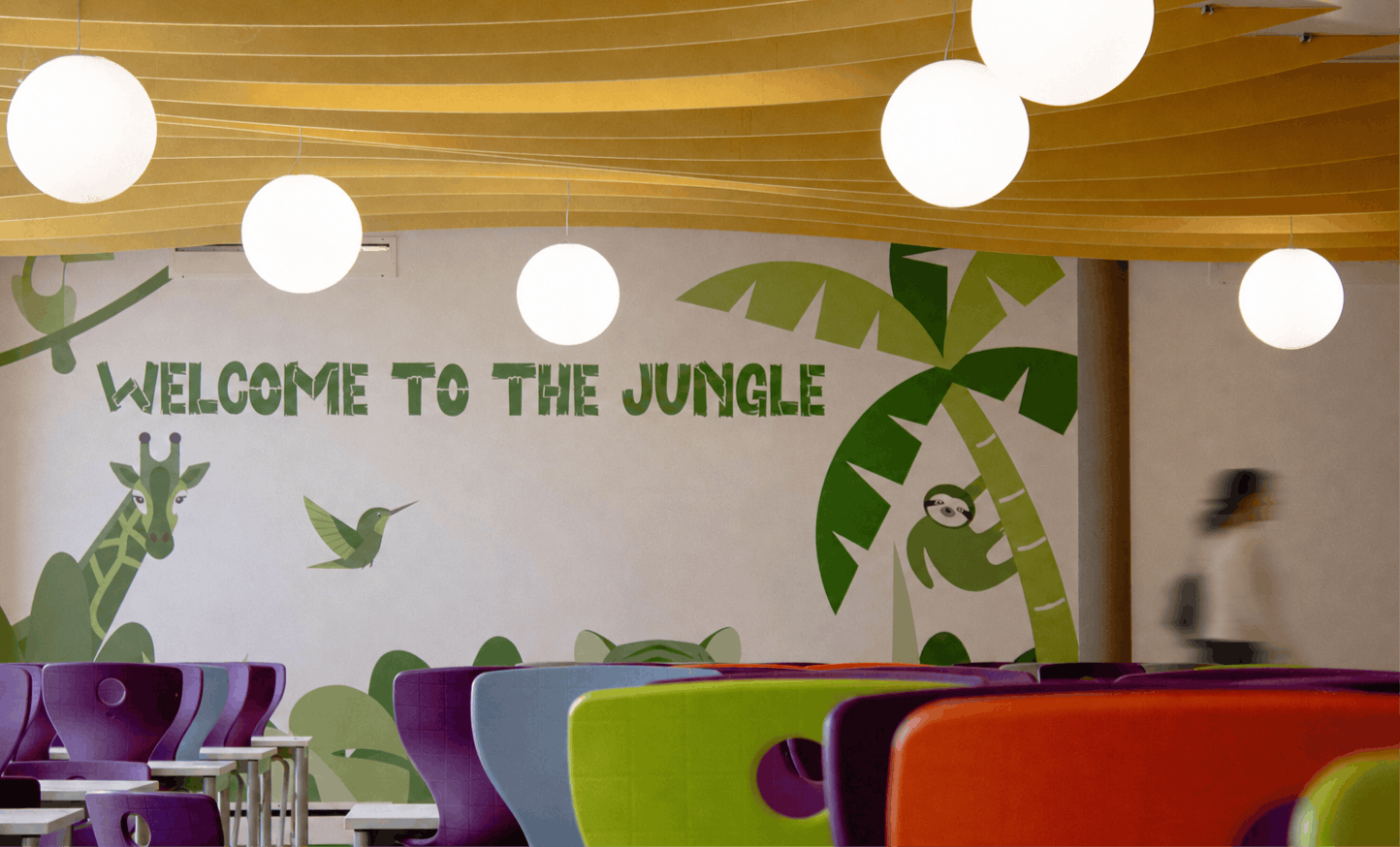 Fresque murale avec l'inscription 'WELCOME TO THE JUNGLE', suspensions lumineuses sphériques blanches et chaises d'école multicolores