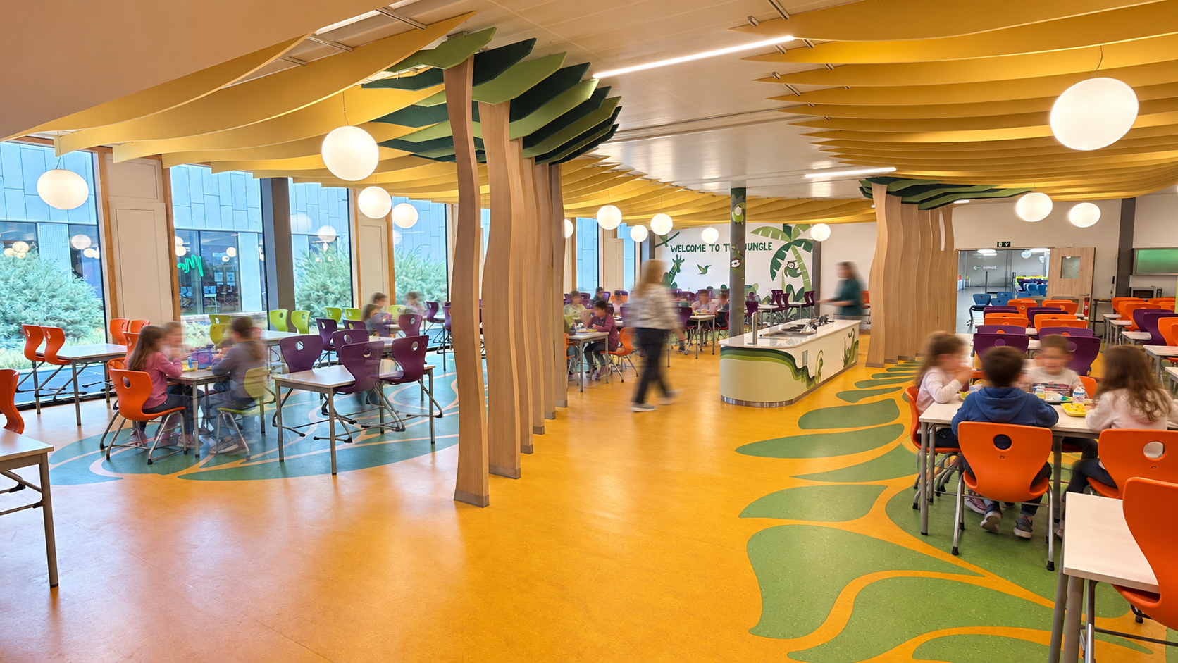 Réfectoire scolaire décoré sur le thème de la jungle avec structures d'arbres en bois, faux feuillages au plafond et tables occupées par des élèves